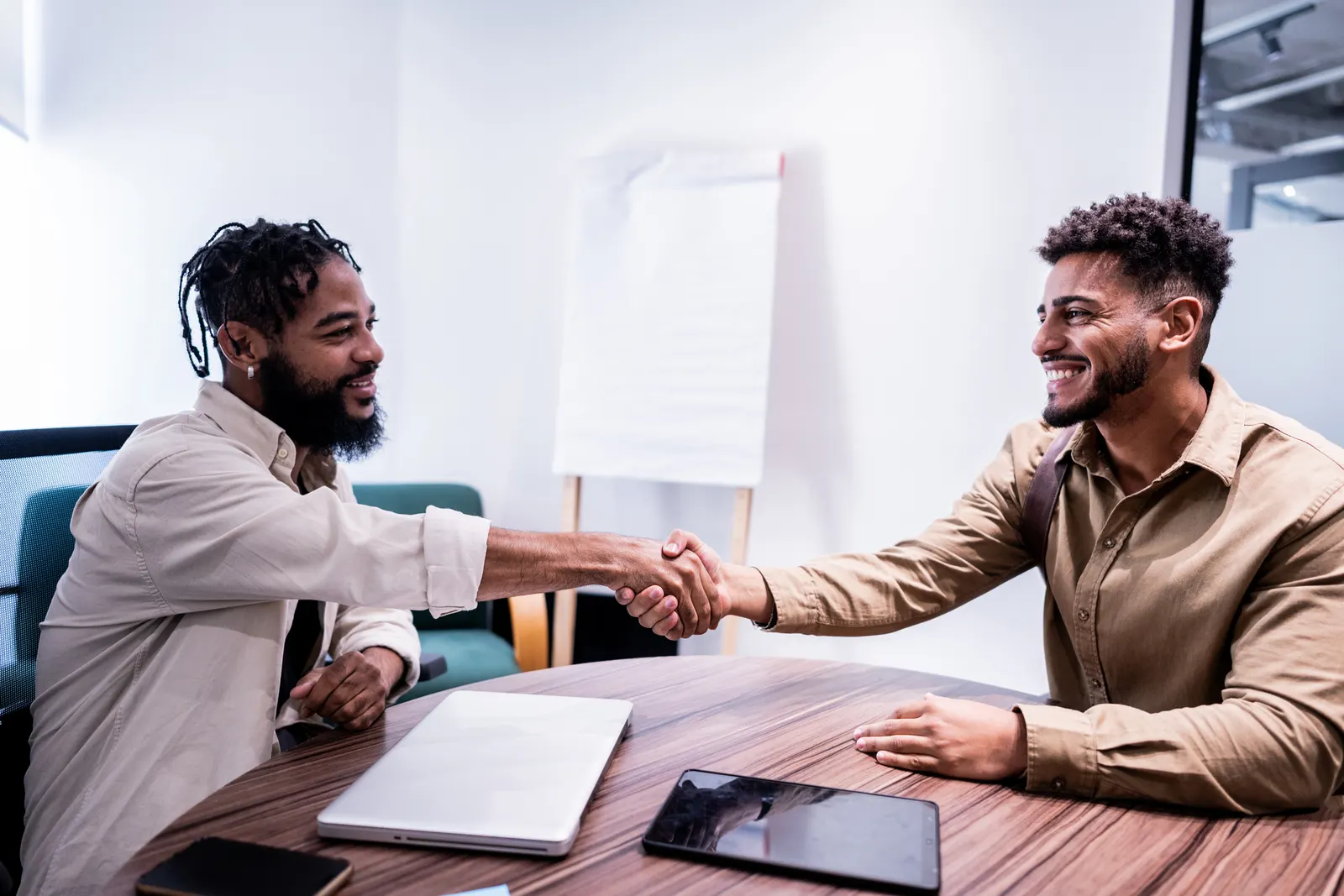 Imagem de dois homens no emprego sentados em frente a uma mesa de cumprimentando com um aperto de mão enquanto se olham e sorriem.