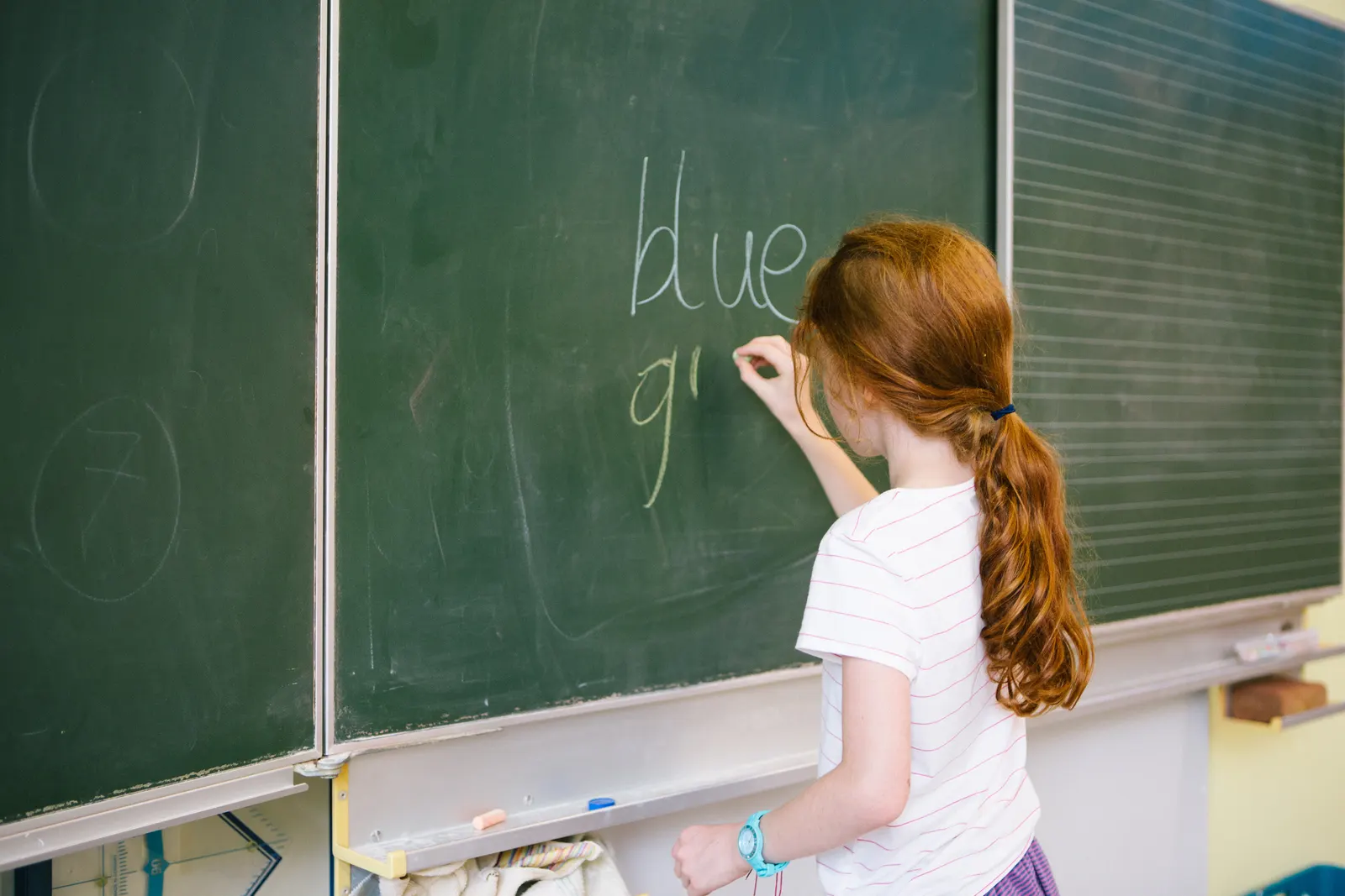 Imagem de um curso de inglês para criança com uma menina de costas escrevendo em inglês duas palavras em uma lousa de giz.