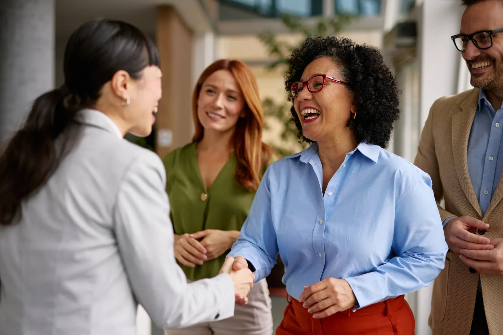 Imagem de duas mulheres sorrindo e se cumprimentando em inglês, ao redor mais uma mulher e um homem olhando elas e sorrindo.