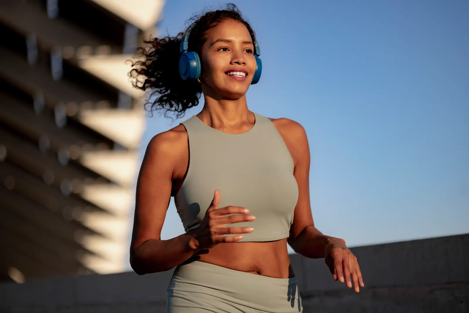 Imagem de uma mulher com conjunto cinza de academia, cabelo preso e fones de ouvido, correndo na rua com uma estrutura desfocada ao fundo. A cena ilustra o estrangeirismo 'Fitness'.