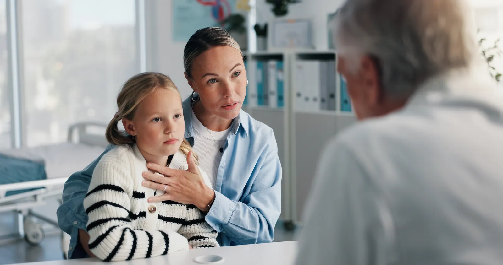 Pronomes pessoais em espanhol. Imagem da mãe sentada em uma cadeira de um consultório com a filha pequena no colo em frente a um médico que está de costas para a imagem desfocada nele.