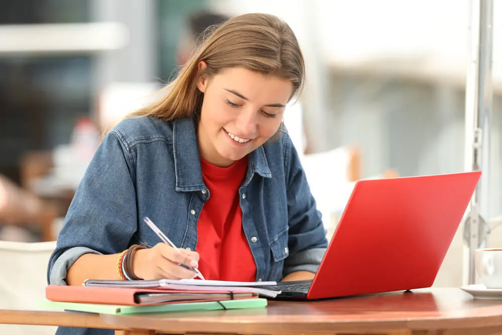 Menina estudando em mesa usando um notebook e anotando em um caderno