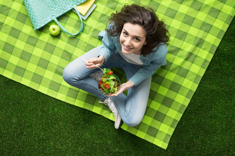 Menina comendo salada sentada em cima de uma toalha estendida sobre um gramado