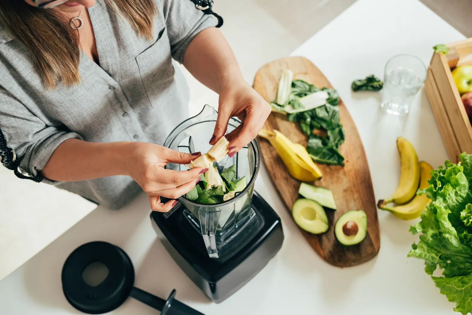 Imagem tirada de cima de uma mulher em frente a uma bancada, colocando uma fruta dentro de um liquidificador, com uma tábua e outras frutas ao redor.