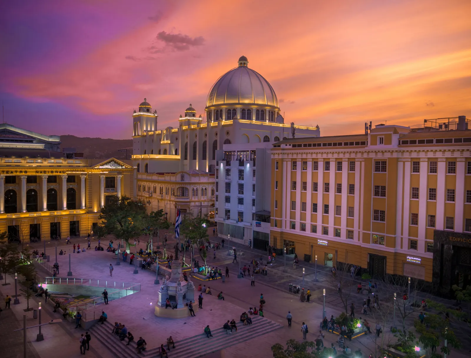 Imagem de um edifício histórico em El Salvador, parcialmente iluminado, com uma praça à frente e pessoas andando; ao fundo, um céu de fim de tarde.