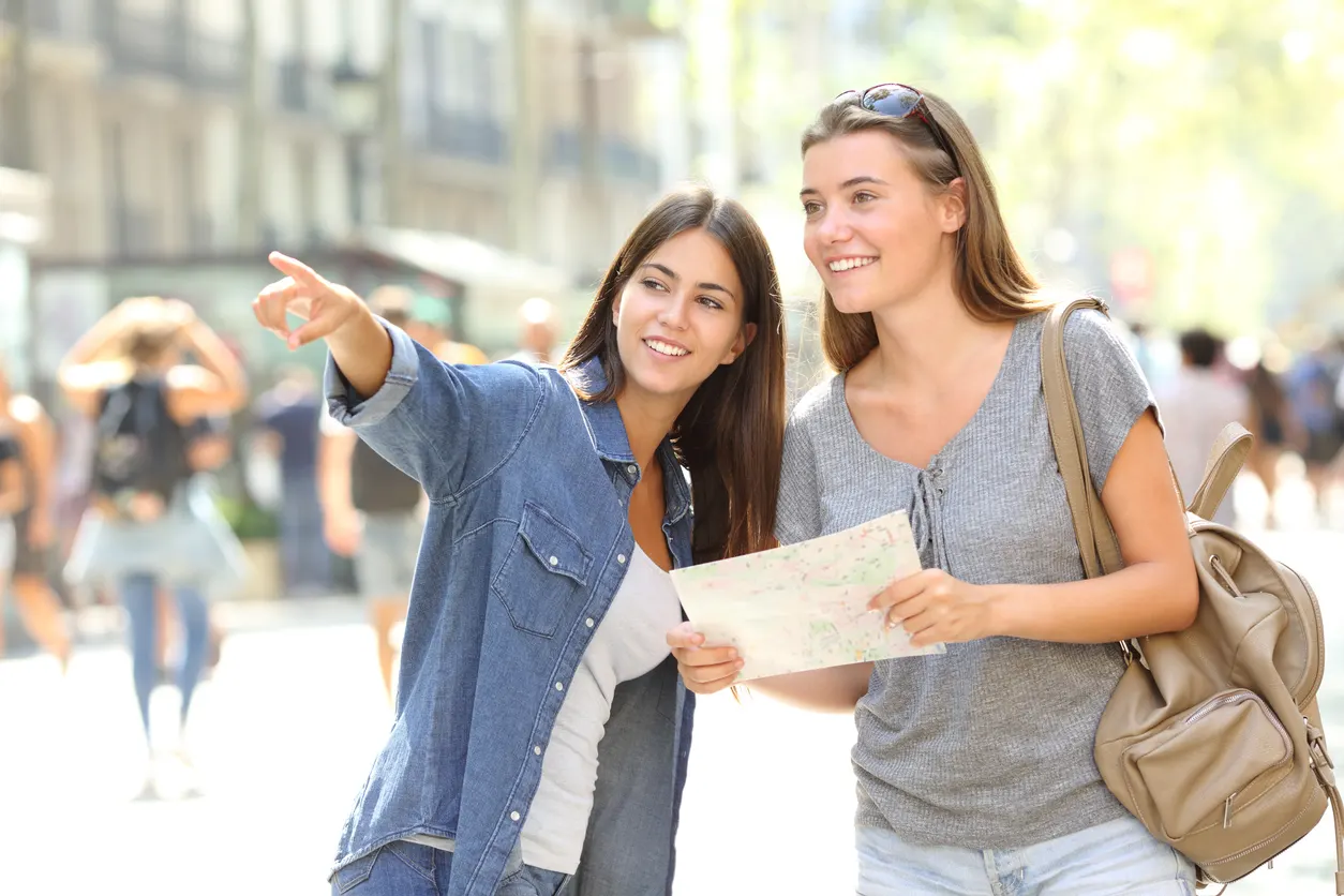 Duas mulheres jovens em uma rua movimentada, uma segurando um mapa e a outra apontando para a direita.