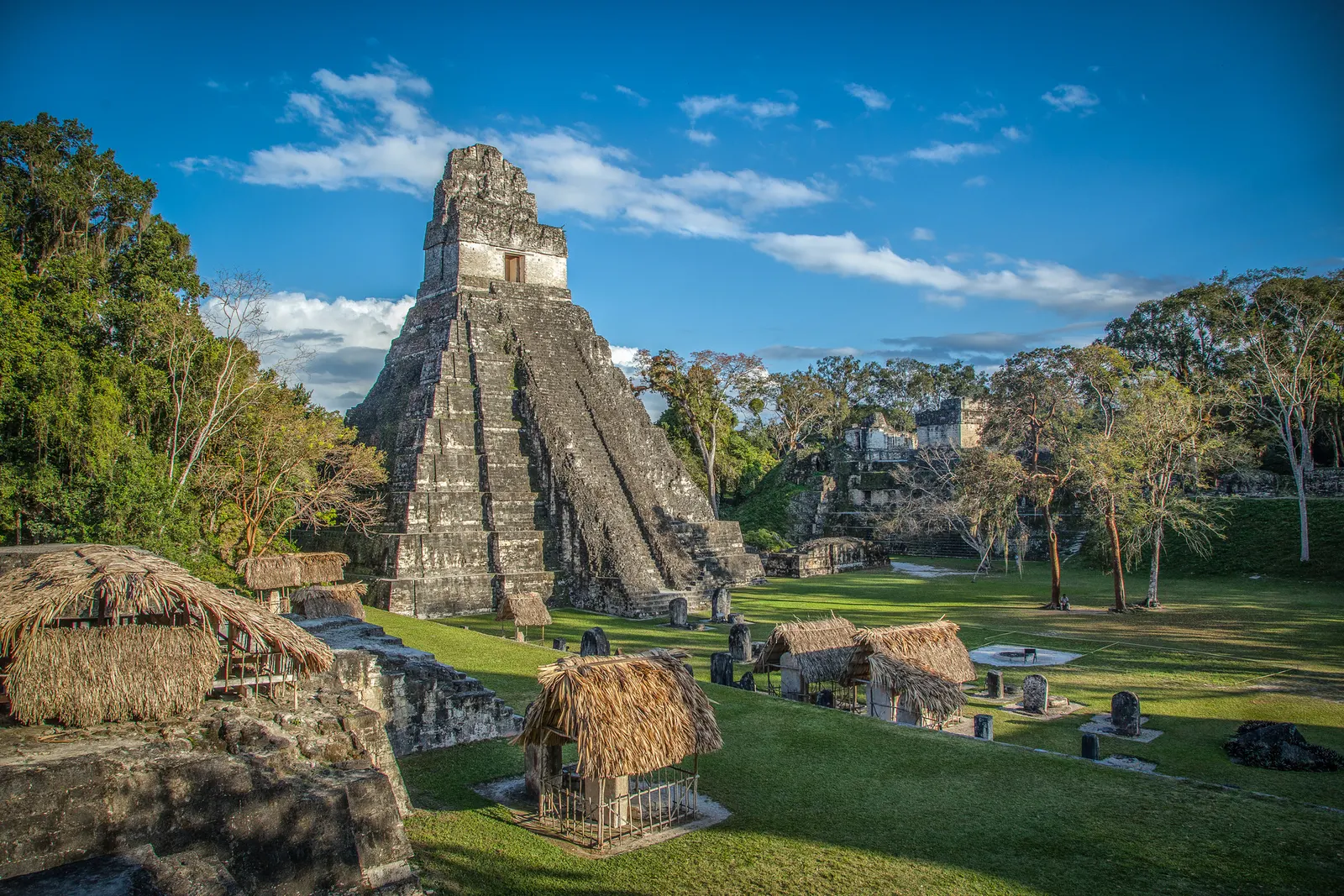 Imagem de uma ruína arqueológica na Guatemala, vista de longe, rodeada por grama e árvores, com pessoas andando ao seu redor.