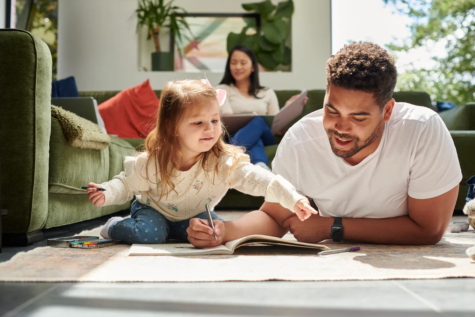 Imagem de uma pai com a filha deitados no chão da sala estudando inglês com um sofá na lateral e outro no fundo com uma mulher sentada.