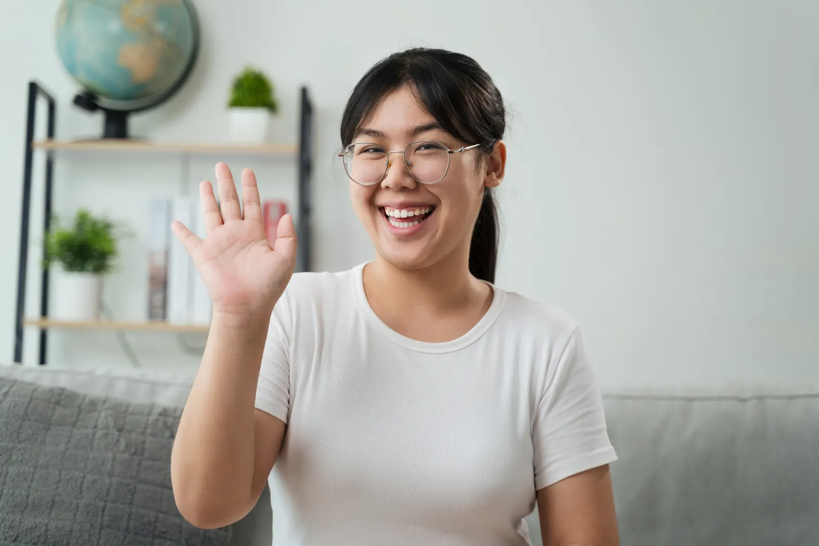 Imaem de uma mulher sentada em um sofá sorrindo com a mão pro alto acenando dizendo "oi" em inglês e uma estante com decoração nos fundos.