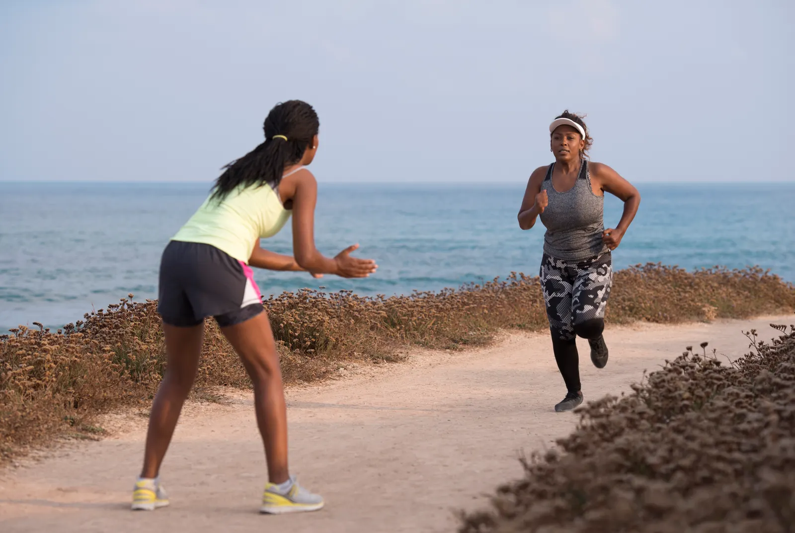 Com o mar ao fundo, uma personal trainer de roupa de academia incentiva com palmas uma mulher que corre à sua frente. A corredora usa o mesmo tipo de roupa e uma viseira. A cena representa o estrangeirismo 'Personal Trainer'.