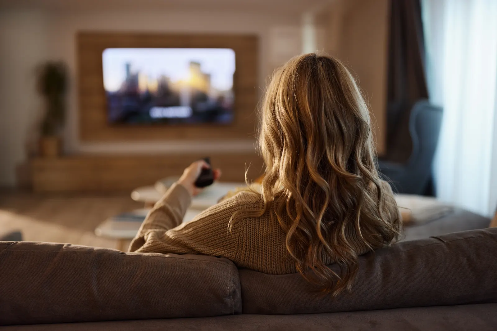 Imagem de uma mulher vista de costas sentada em um sofá apontando o controle na direção da TV, enquanto assiste um filme em inglês.