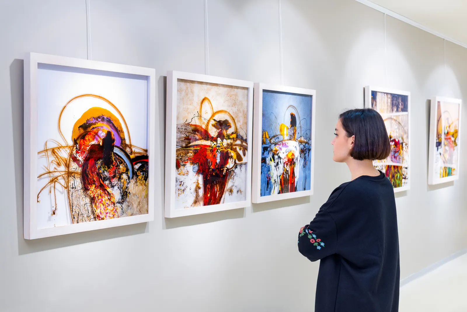 Vista lateral de uma jovem mulher com cabelo curto e escuro, vestindo uma blusa escura, admirando uma série de quadros abstratos vibrantes pendurados em uma parede clara de uma galeria de arte.