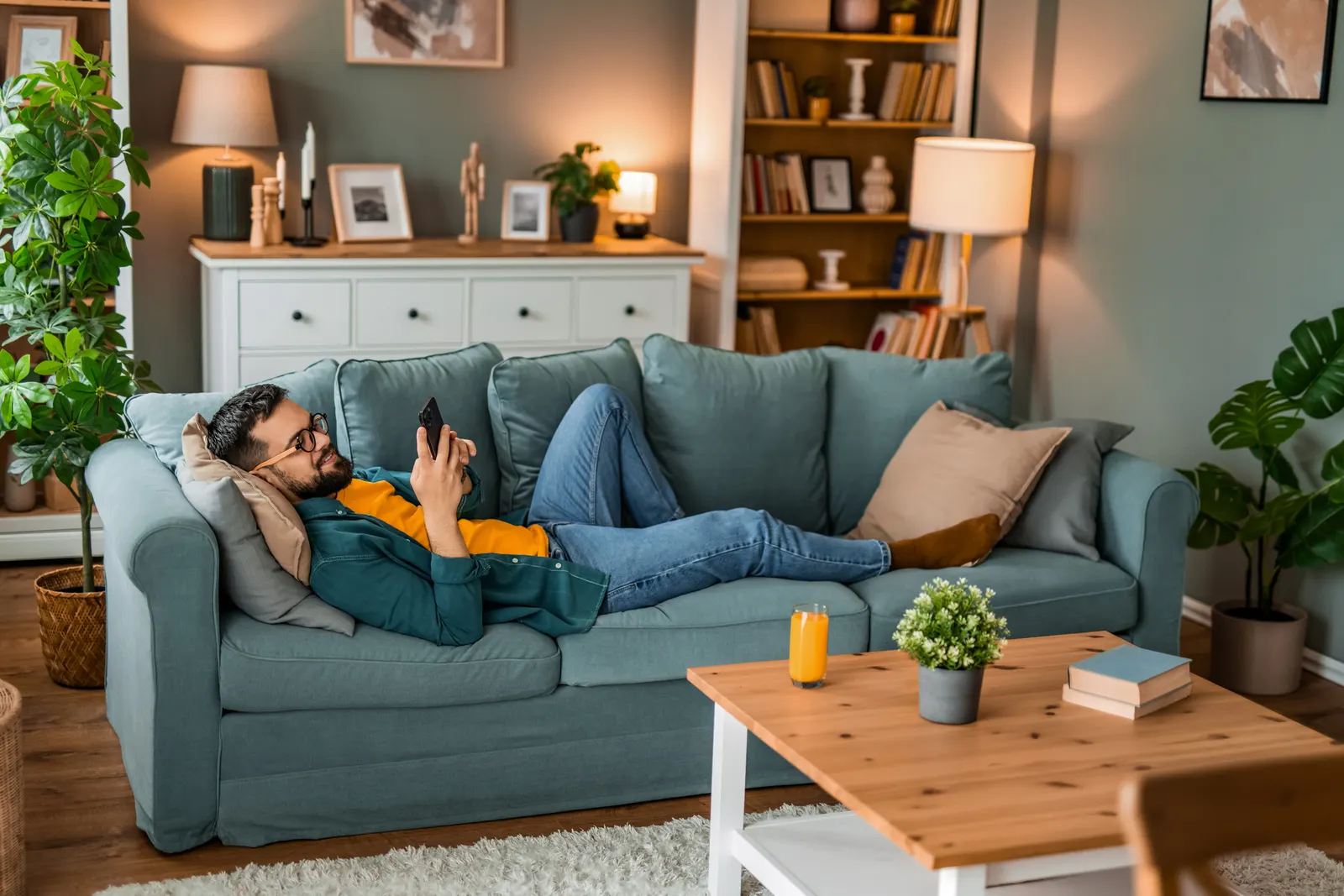 Imagem de um homem mexendo no celular enquanto está deitado em um sofá azul em uma sala com uma mesa de centro de madeira na frente e armários aos fundos.