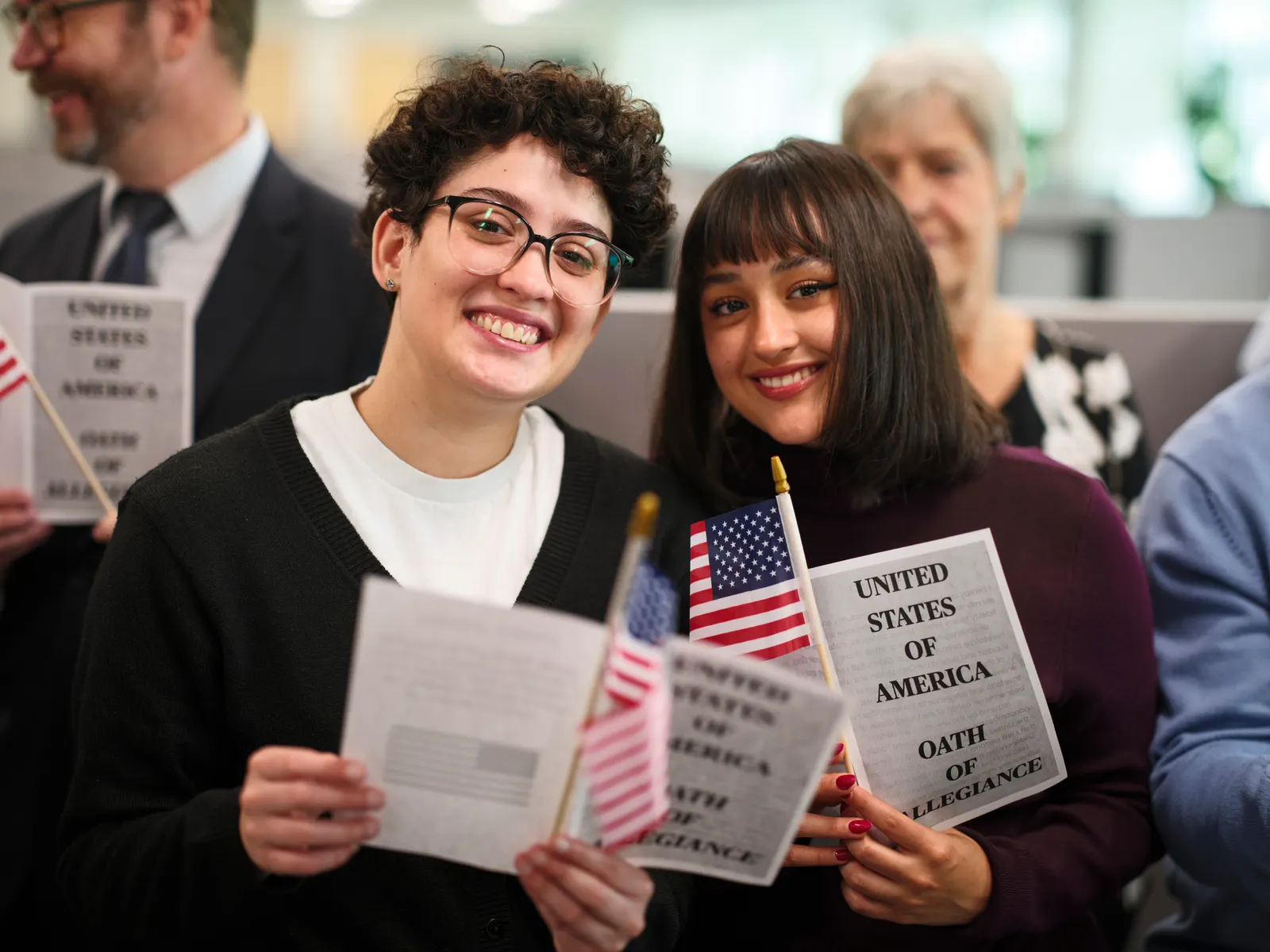 Imagem de duas pessoas uma do lado da outra sorrindo enquanto olham para a câmera, cada uma segurando uma bandeira pequena dos Estados Unidos e um folder de como conseguir a cidadania americana.