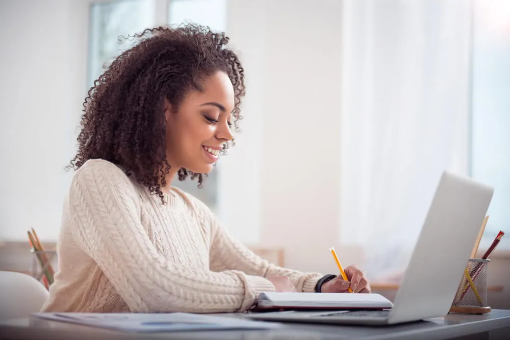 Mulher escrevendo em caderno em frente a um computador