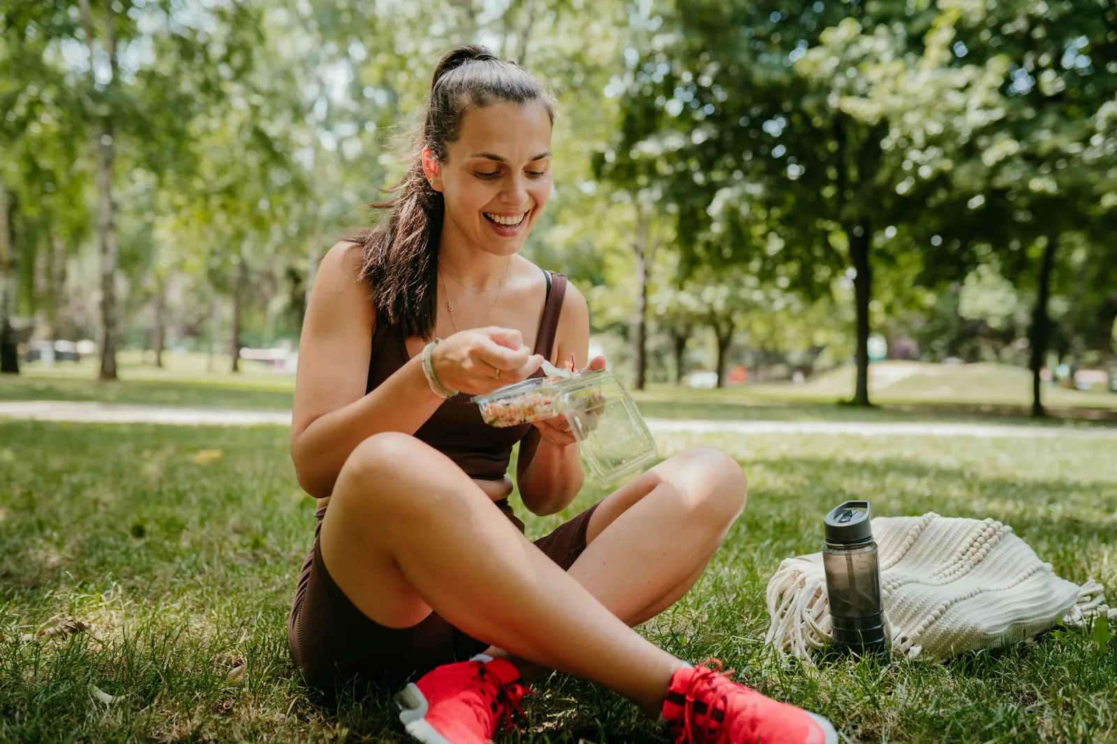 Imagem de uma mulher com roupa de academia, sentada na grama de um parque com árvores ao fundo. Ela segura um pote com algum alimento light. A imagem representa o termo de estrangeirismo 'Light'.