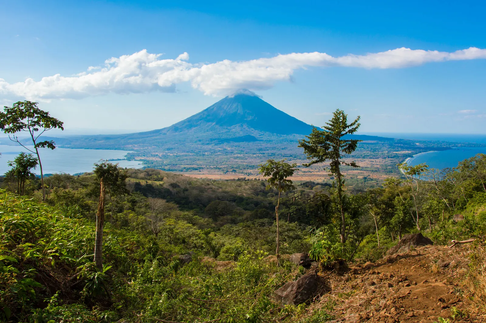 Imagem de uma área verde com plantações; ao fundo, um vulcão sob um céu azul com nuvens e, à frente do vulcão, uma praia.