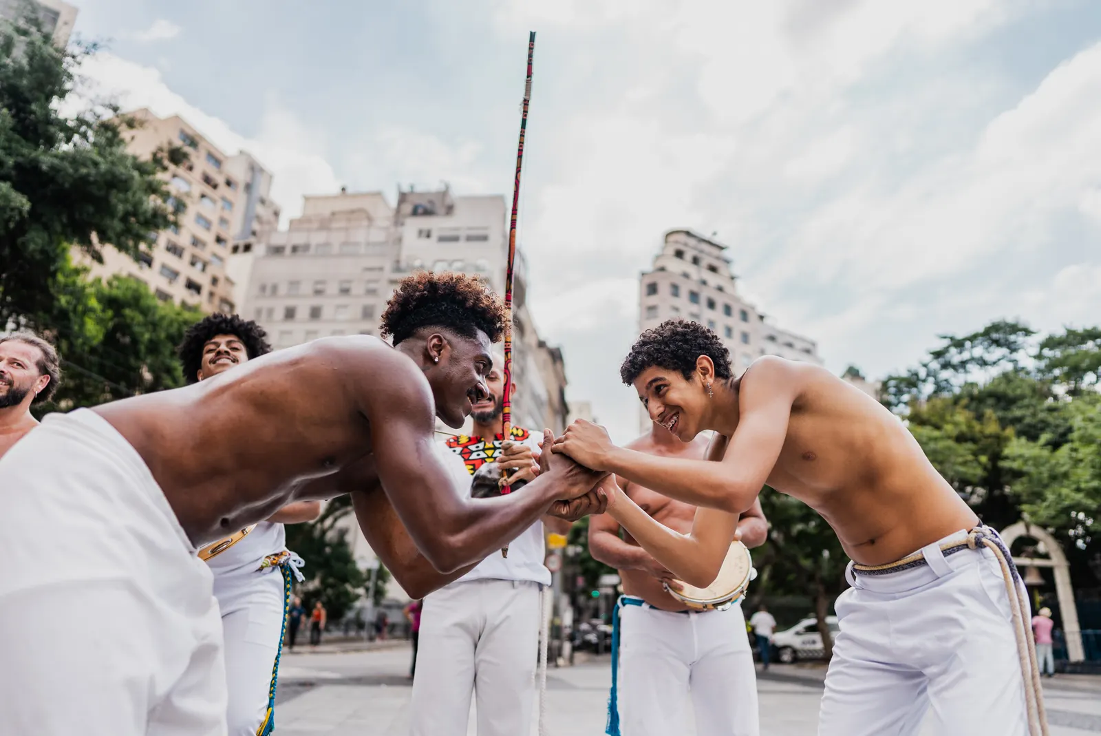 Imagem de vários homens reunidos em uma praça, praticando capoeira, com prédios e árvores ao redor.