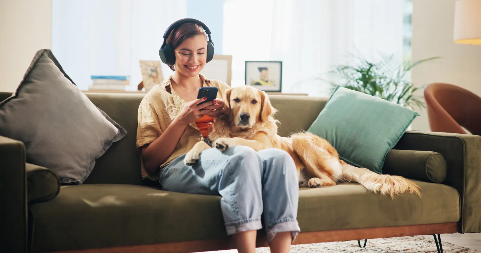 Imagem de uma mulher sentada em um sofá, usando fones de ouvido e com um celular na mão. Ela escuta um podcast em inglês enquanto um cachorro Golden Retriever, deitado ao seu lado, apoia as patas em suas pernas.